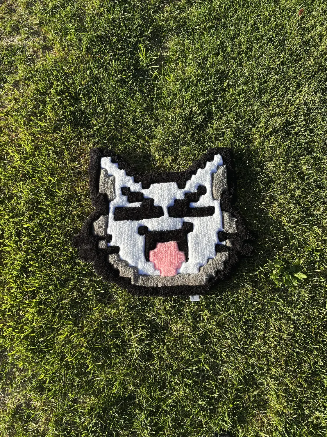 Black, white and grey pixel-style cat face rug with a pink tongue on grass