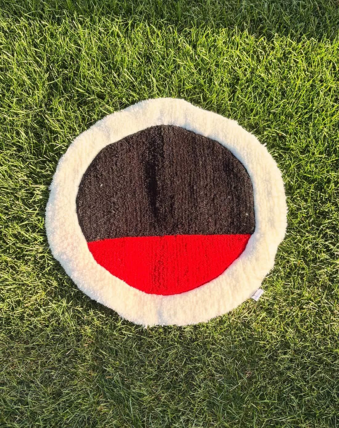 Round hand-tufted rug on grass: black top half, red bottom half, thick white pile edge
